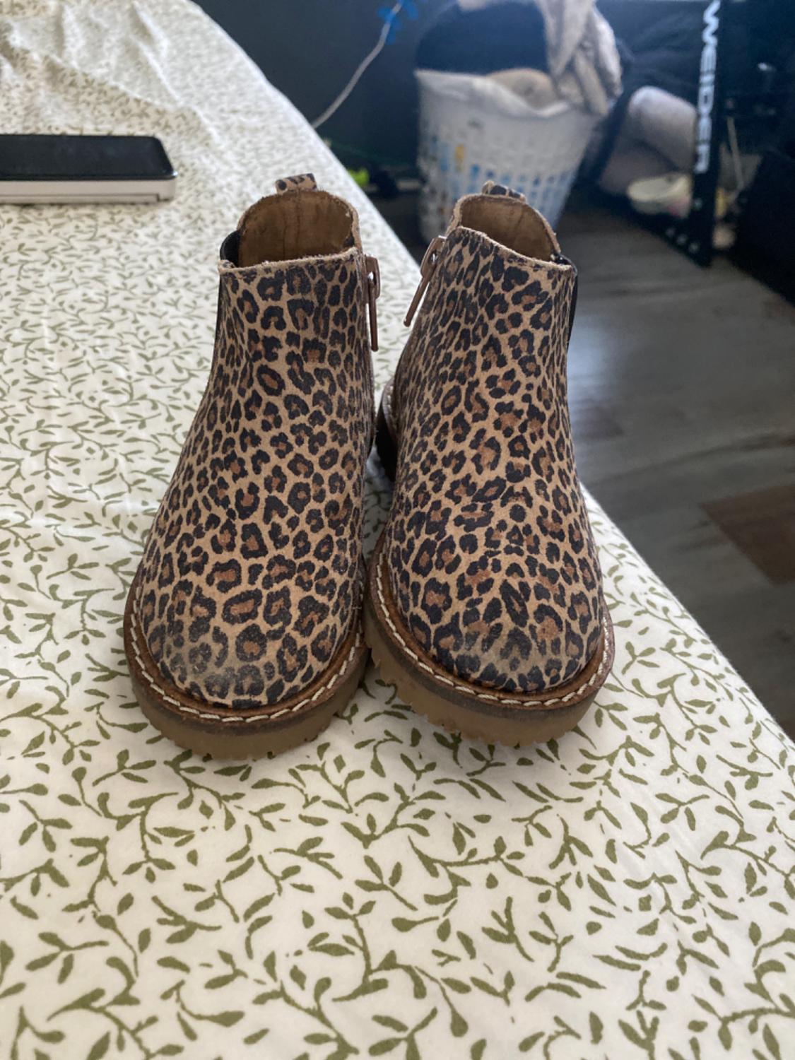 Leopard print ankle boots on a white quilt with a green leaf design, with a laundry basket and household objects in the background.