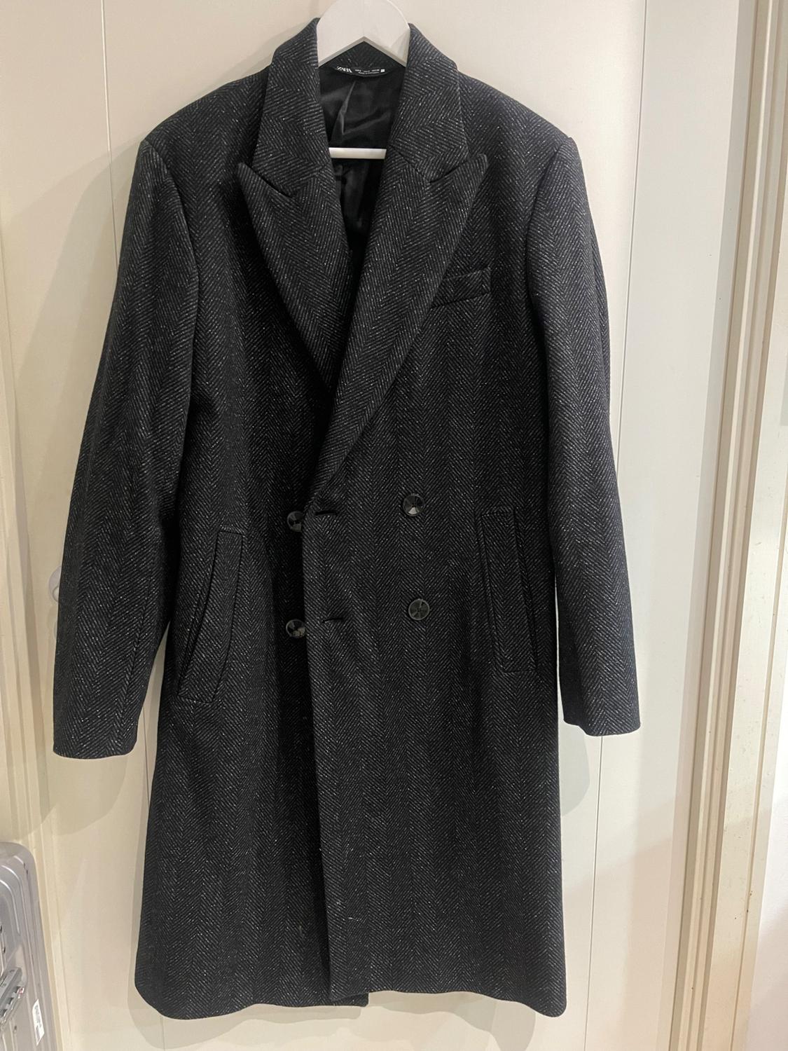 Dark grey long coat with a herringbone pattern, hanging on a white hanger against a light wall.