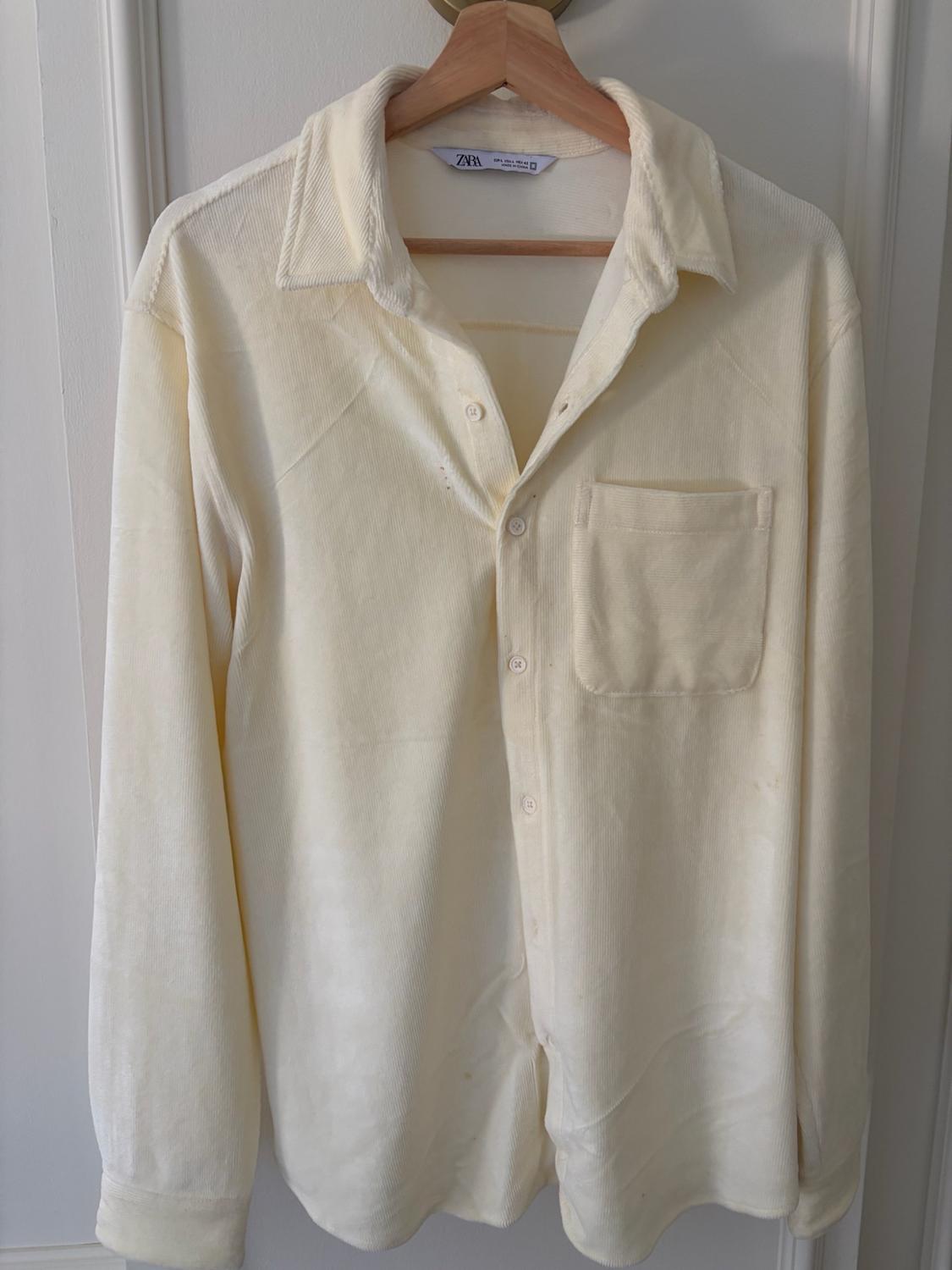 Cream-coloured long-sleeved shirt with buttons and a pocket, hanging on a wooden hanger against a white background. ZARA label on the collar.