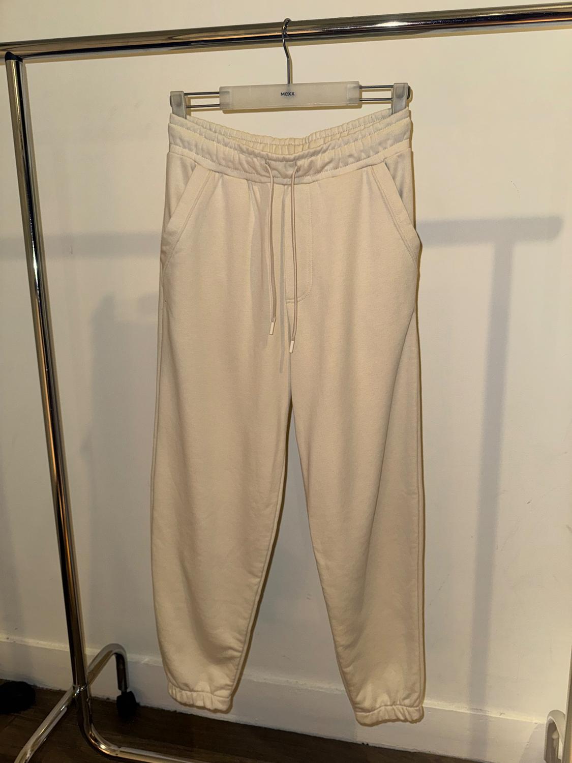 Beige tracksuit bottoms hanging on a metal hanger, with adjustable drawstring and side pockets, on a white background.