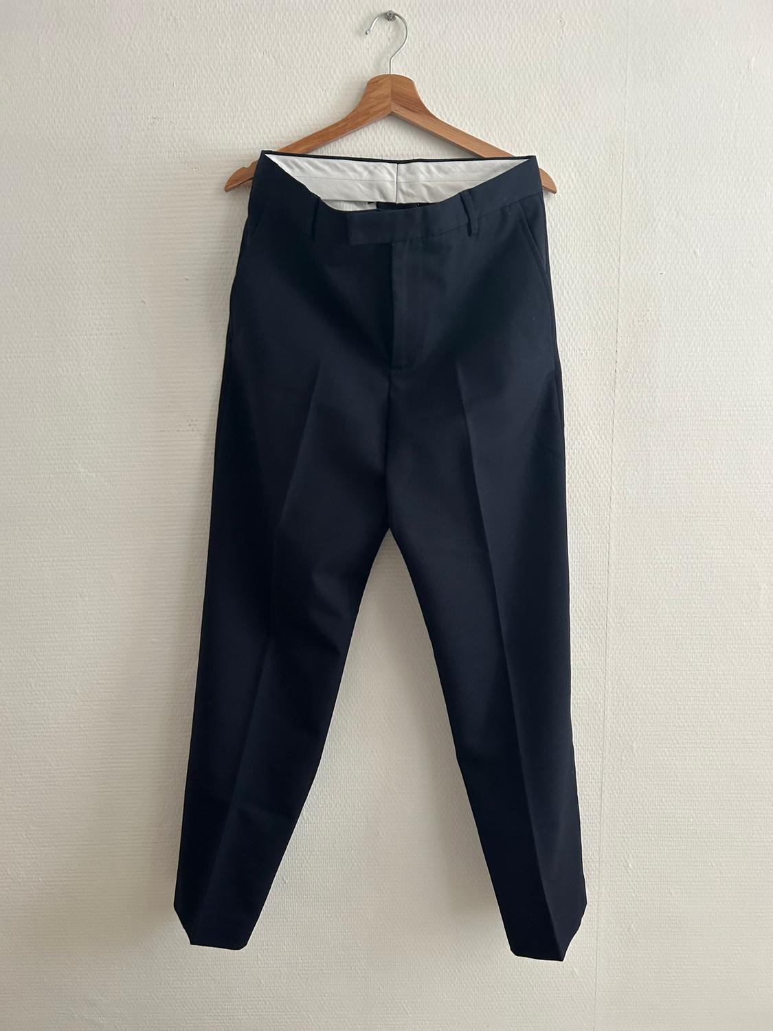 Black trousers hanging on a wooden hanger against a white wall. Classic design with front pleats.