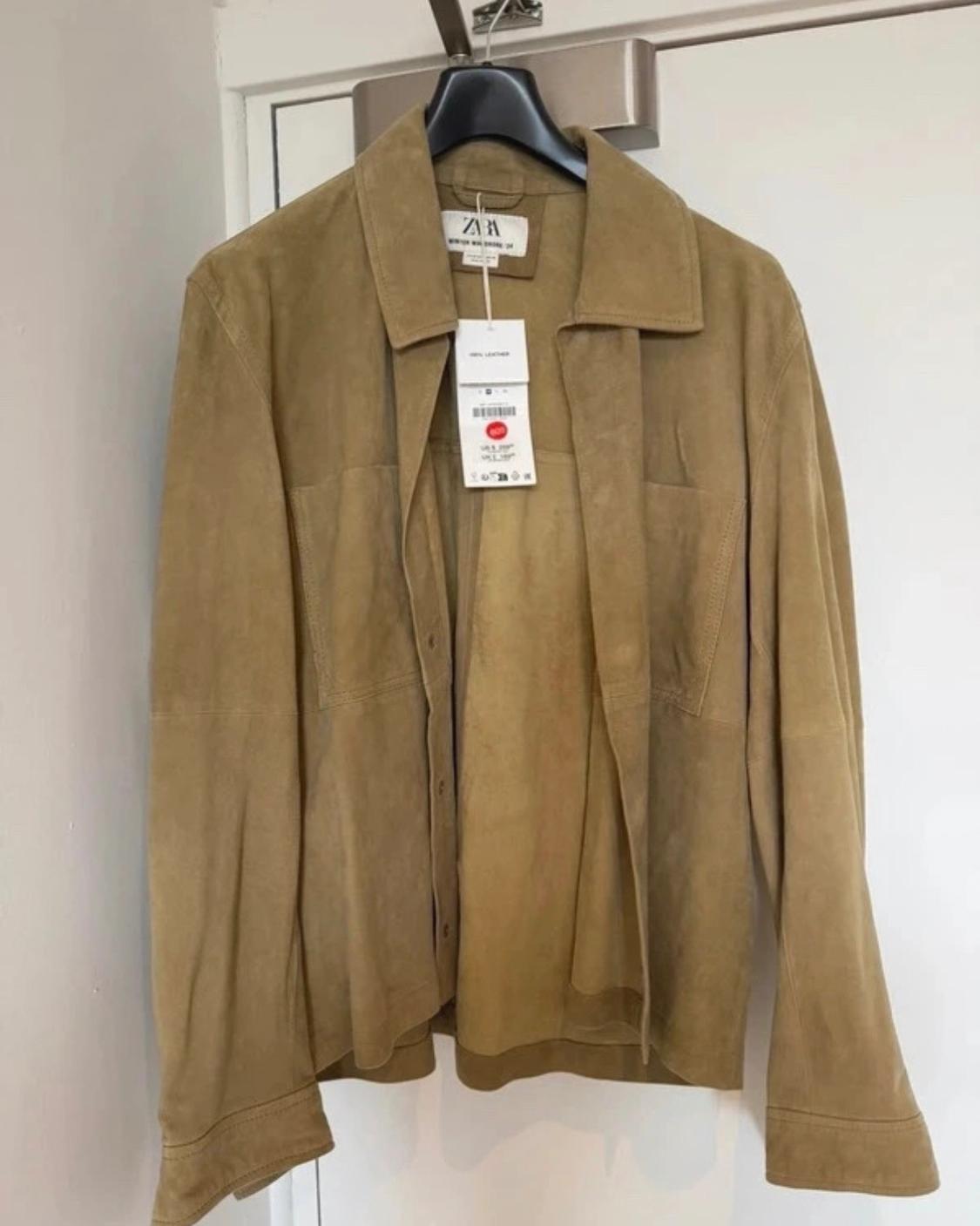 Light brown leather jacket hanging on a hanger on a white door. ZARA label visible with the text 100% LEATHER.