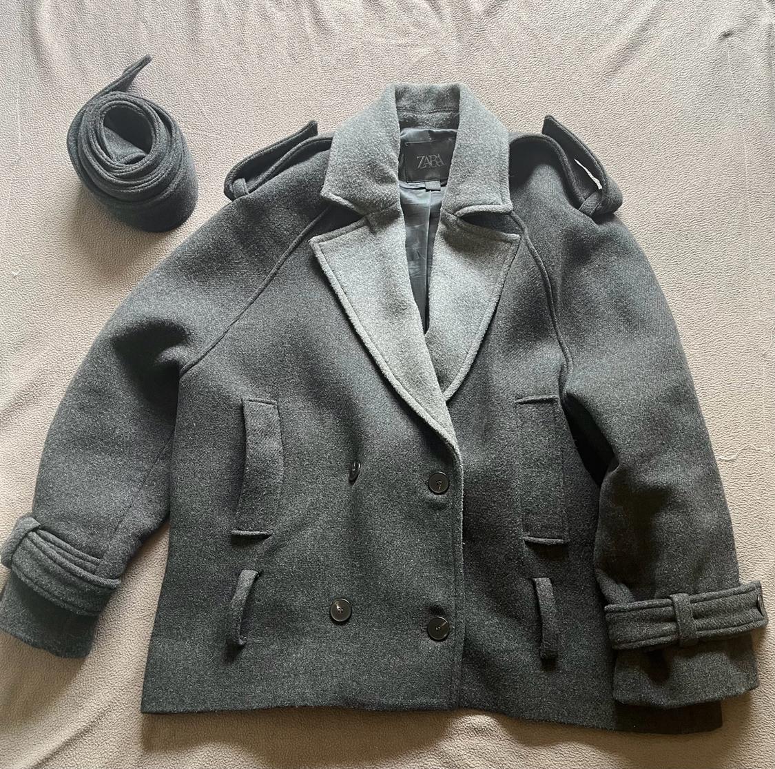 Double-breasted dark grey coat with contrasting lapels and a rolled-up belt at the side, on a fabric surface. Visible text: ZARA.