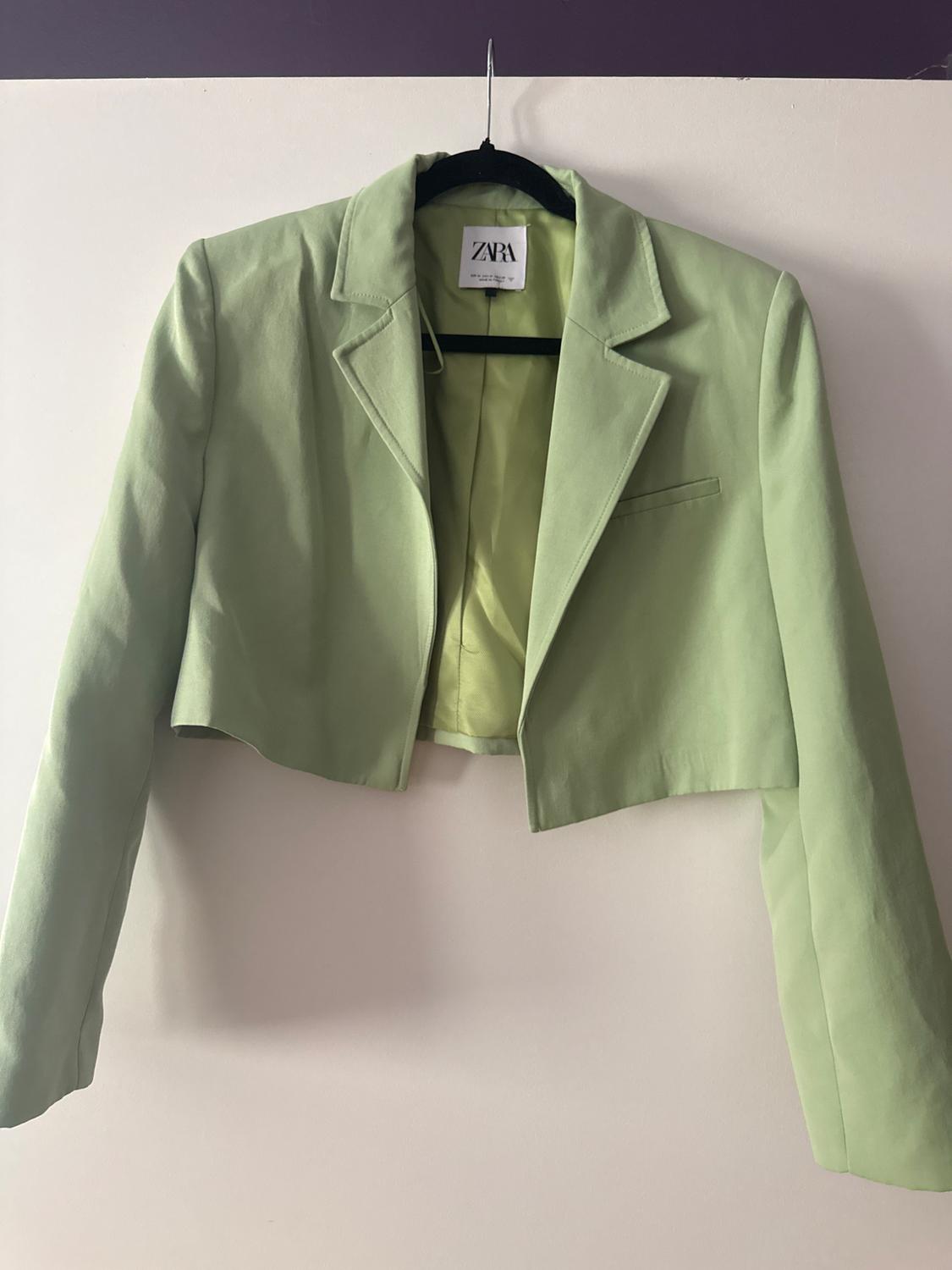 Light green short blazer hanging on a black hanger against a white wall. Inside, a label with the text ZARA is visible.