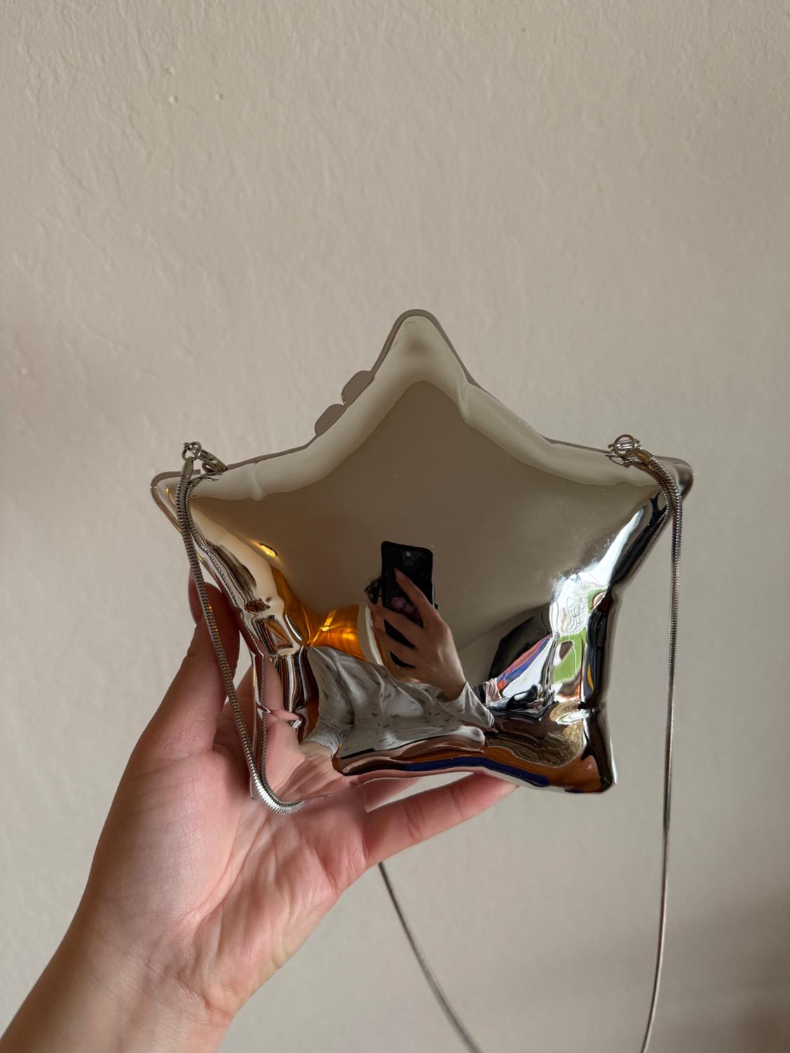 A metallic star-shaped handbag is being held by a hand. A person taking the photo with a mobile phone is reflected on its surface.