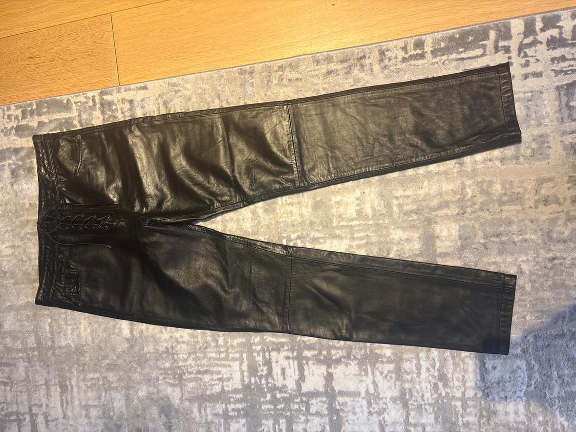 Black leather trousers with embroidered waist details, placed on a grey rug with a geometric pattern.