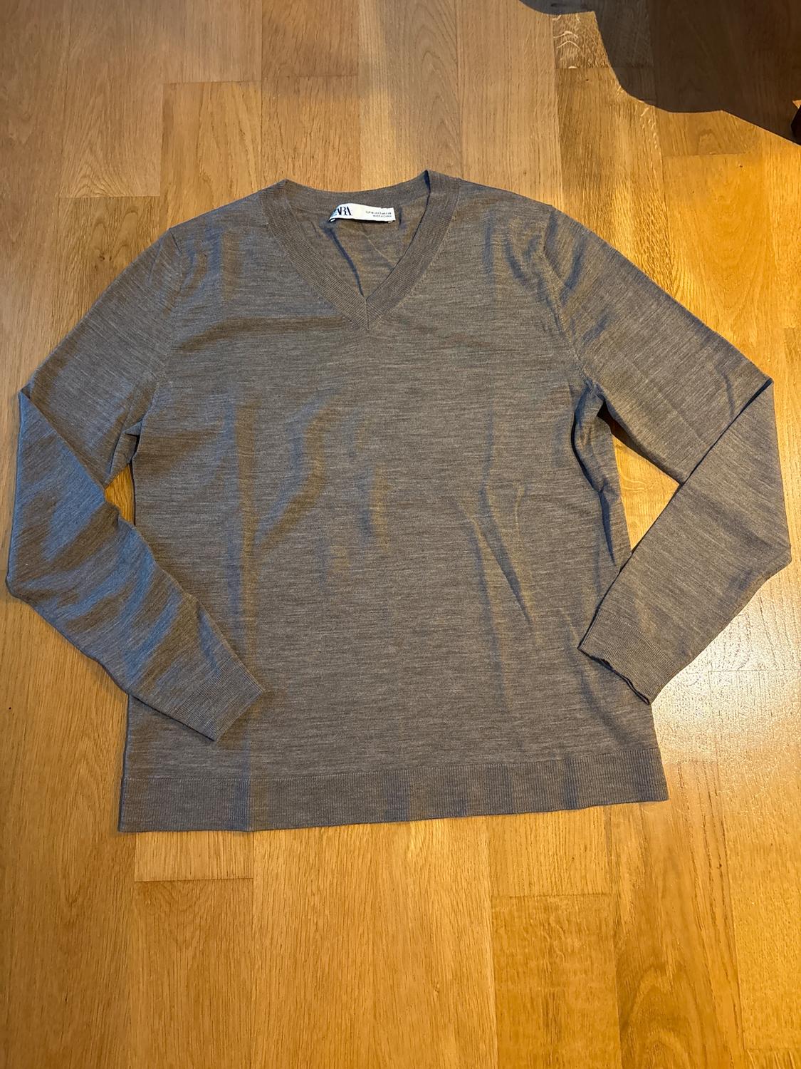 Grey long-sleeved V-neck jumper on a light wooden floor. Visible label on the collar.