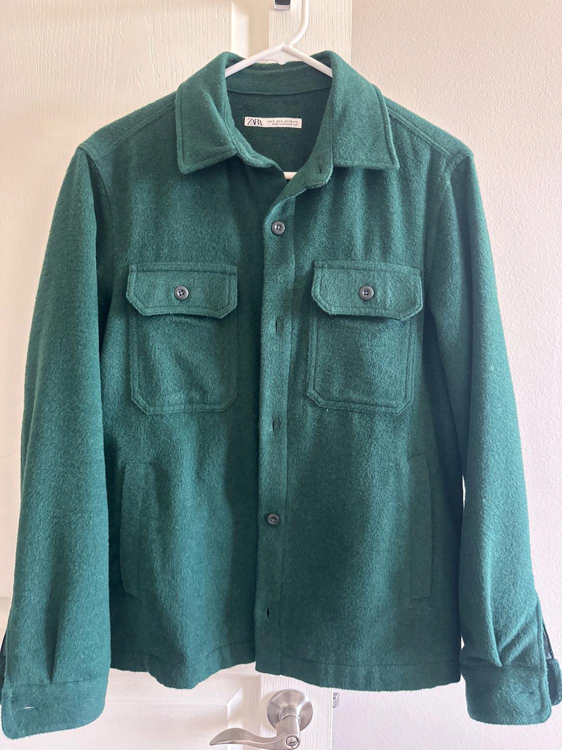 Green wool jacket hanging on a white hanger. It has two flap pockets and buttons on the front.