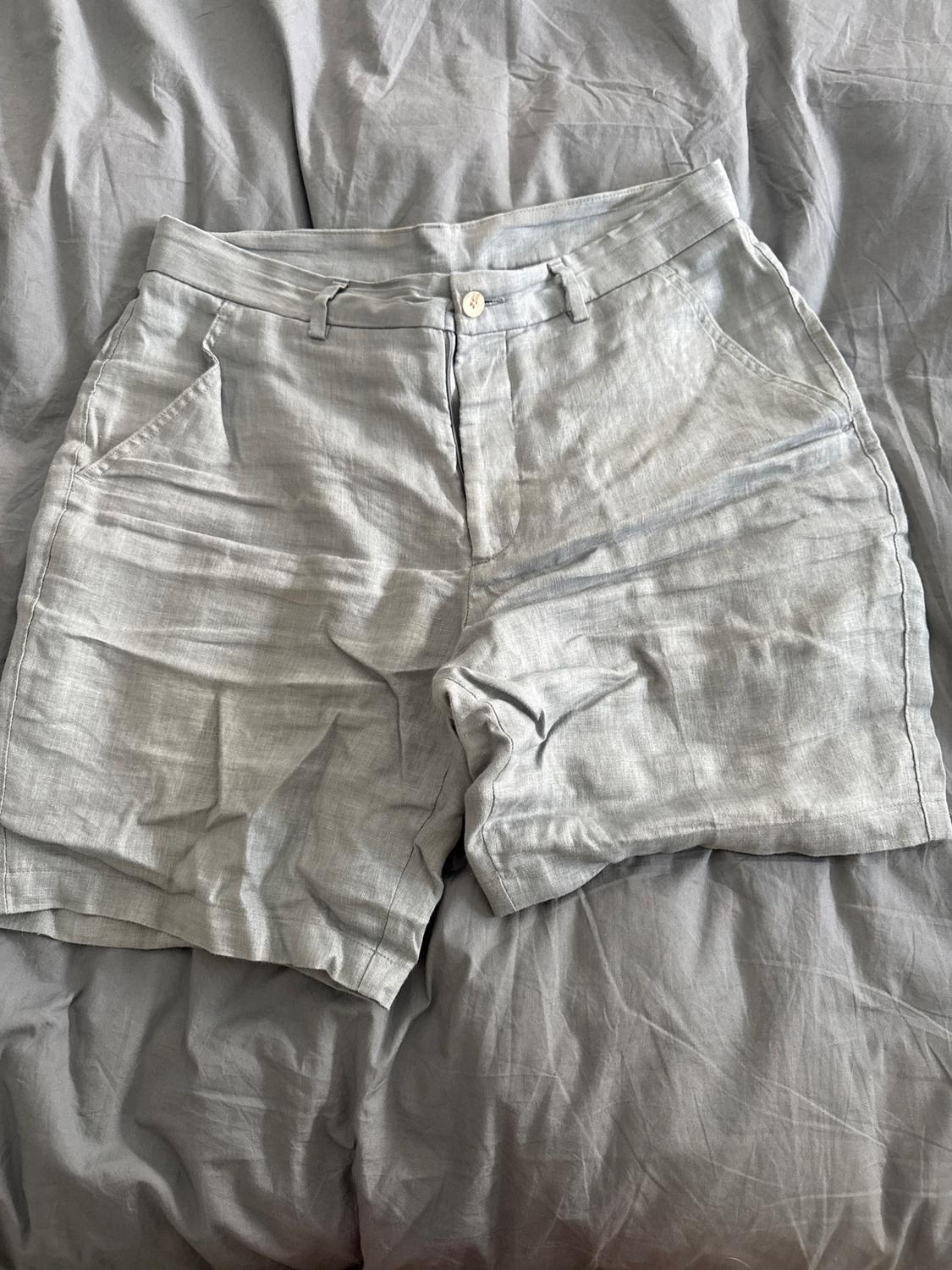 Grey crinkled linen shorts on a grey bed. They have side pockets and a white button fastening.