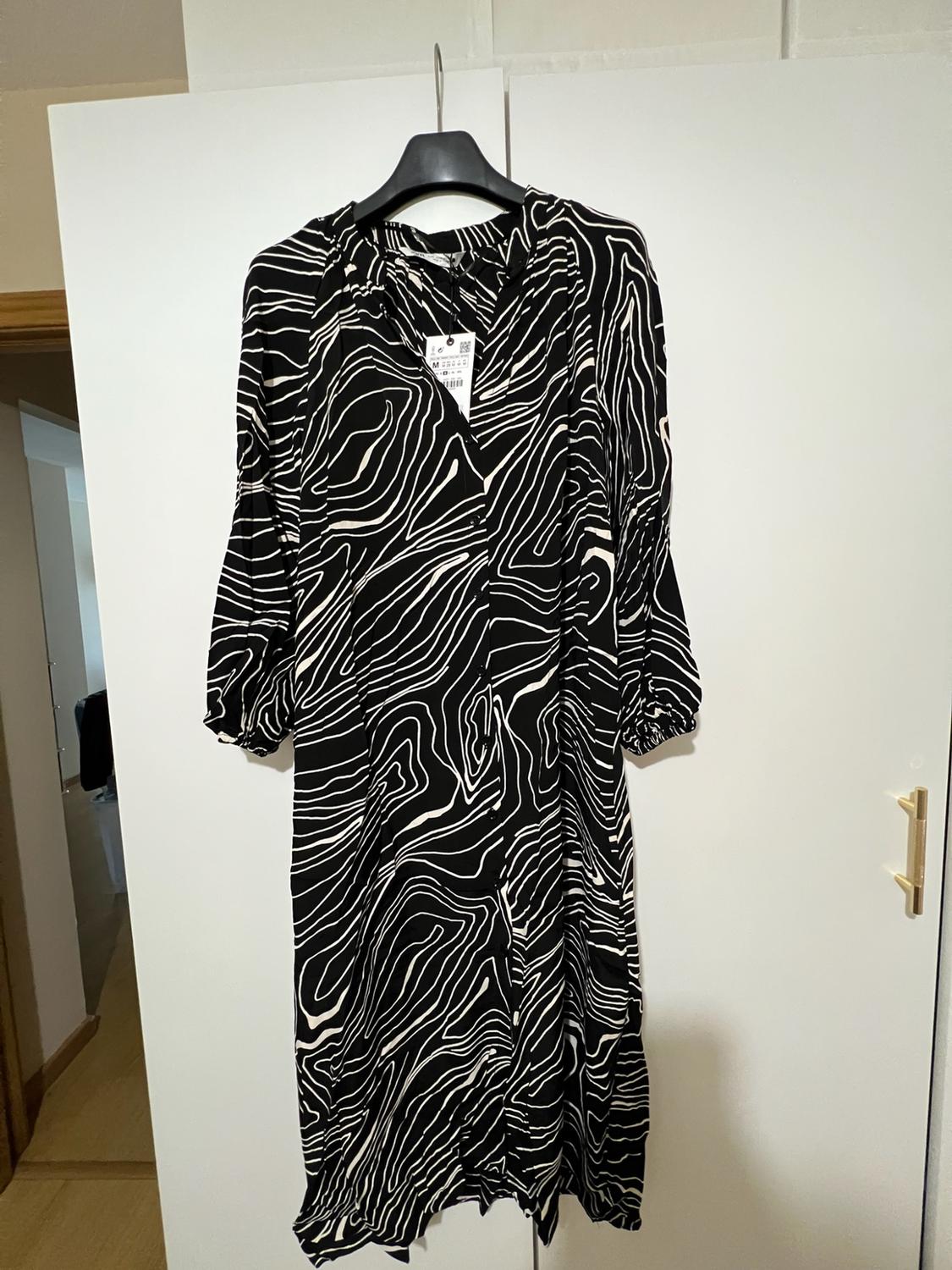 Black dress with a white wavy-line pattern, long sleeves and front buttons, hanging in a white wardrobe.