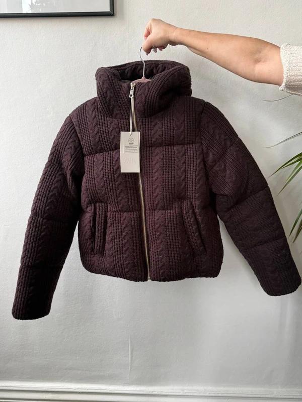 A dark brown quilted jacket is hanging on a hanger and being held by a hand. The label is visible in the centre. The background is white.