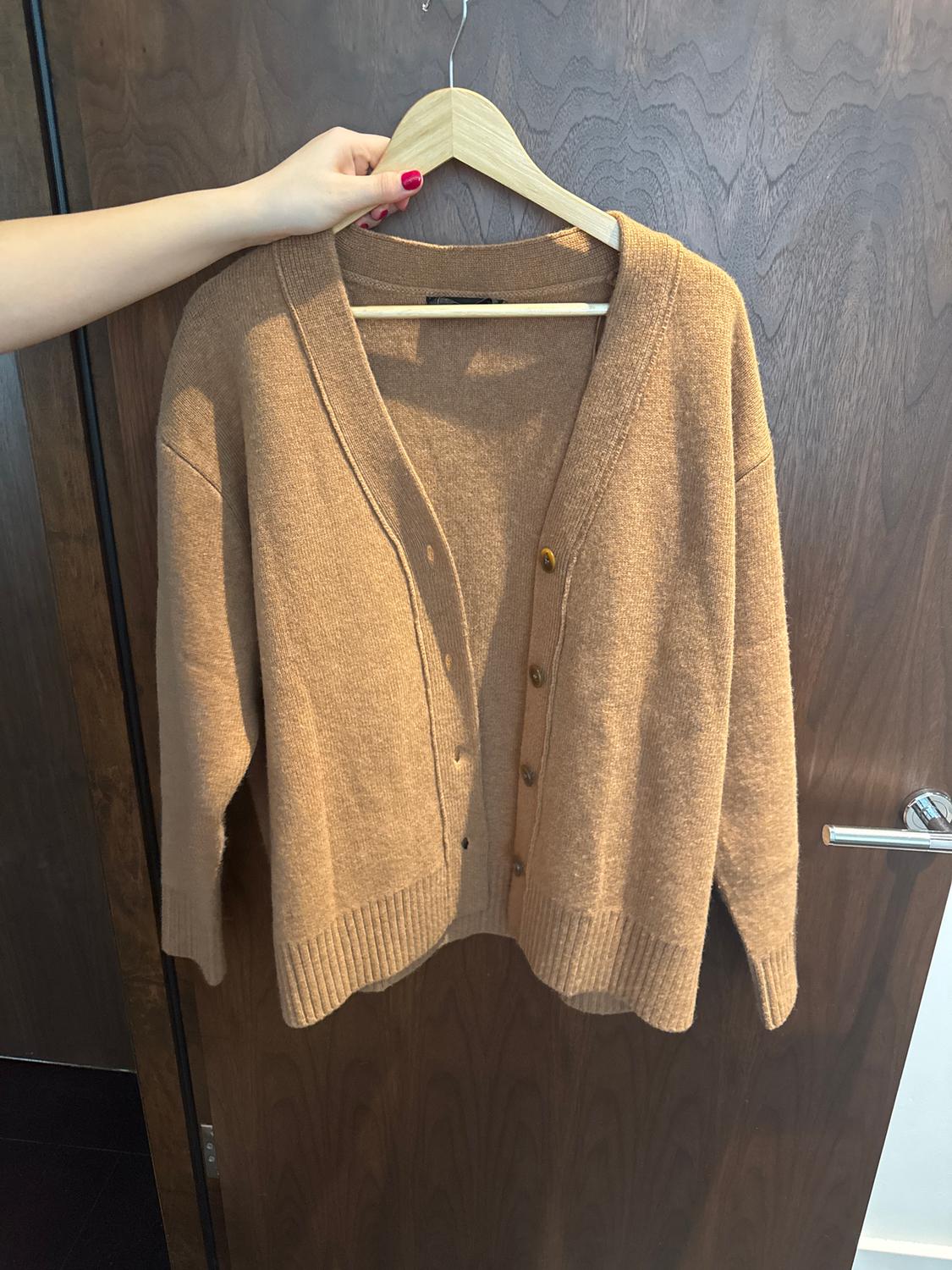 A brown cardigan is hanging on a wooden hanger, held by a hand with red-painted nails, with a wooden door as a background.