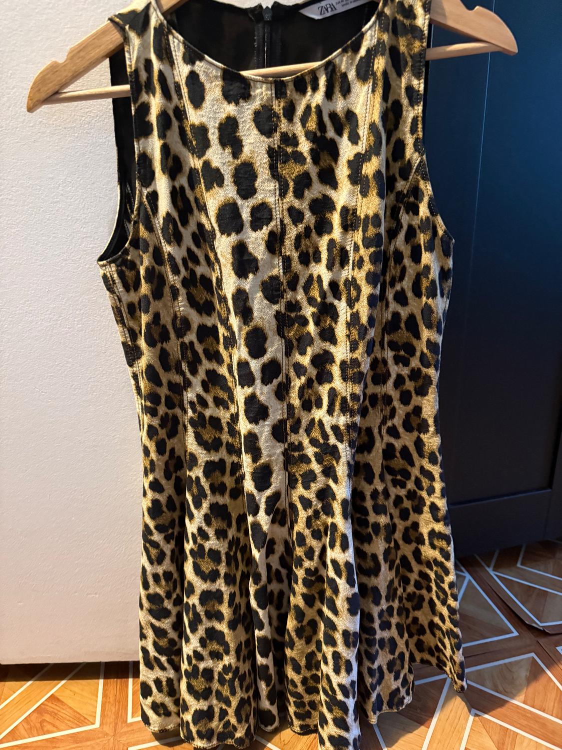 Sleeveless leopard print dress hanging on a wooden hanger. White wall background and wooden floor.