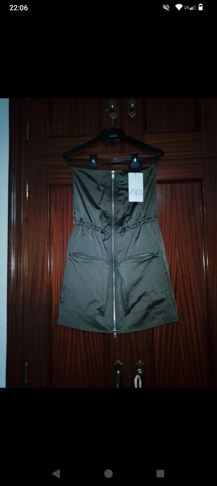 Olive green strapless dress with front zip and pockets, hanging on a black hanger in front of a wooden door. Label visible.