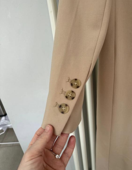 Hand holding the sleeve of a beige coat with three brown buttons. White and light grey background.