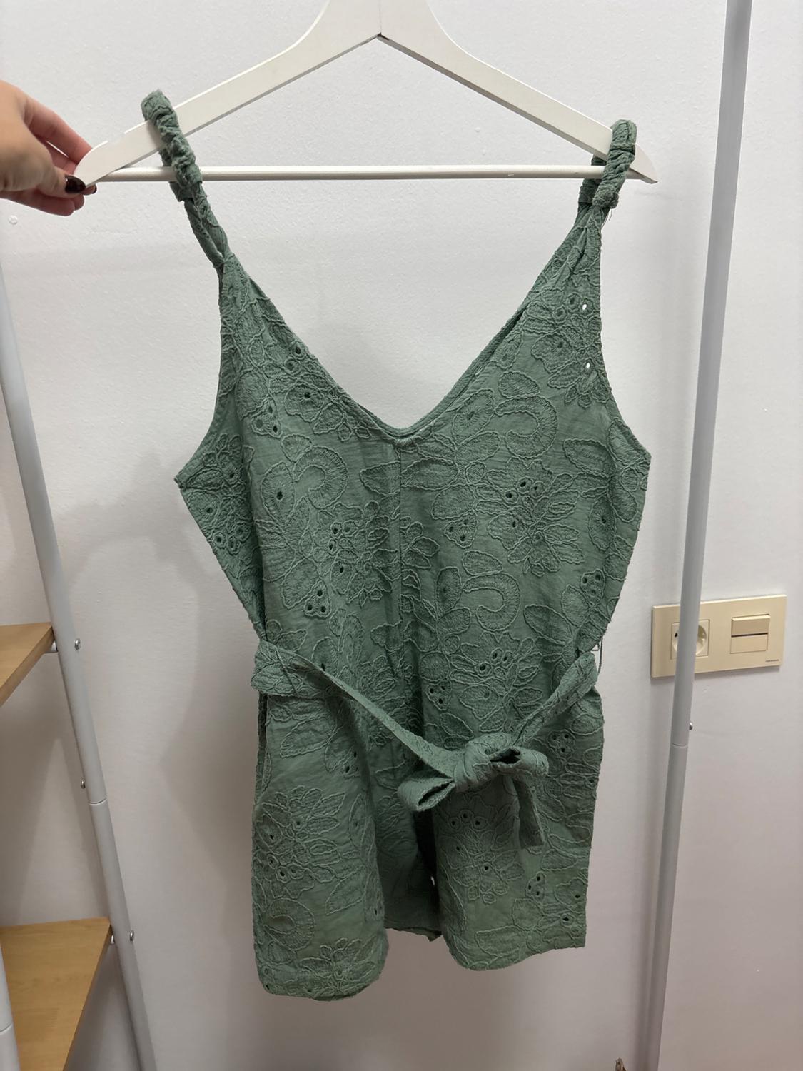 Green jumpsuit with straps and belt, hanging on a white hanger. White background with light switch on the right.