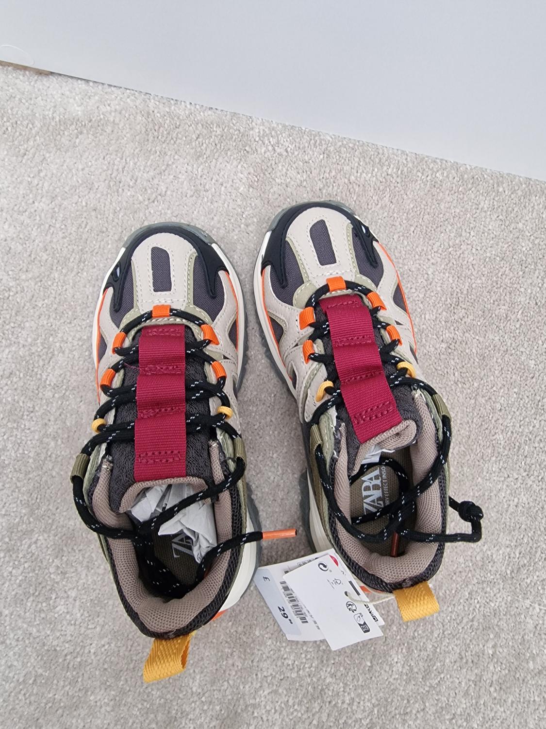Multi-coloured trainers with orange and burgundy detailing, black laces and visible labels on a grey carpet.