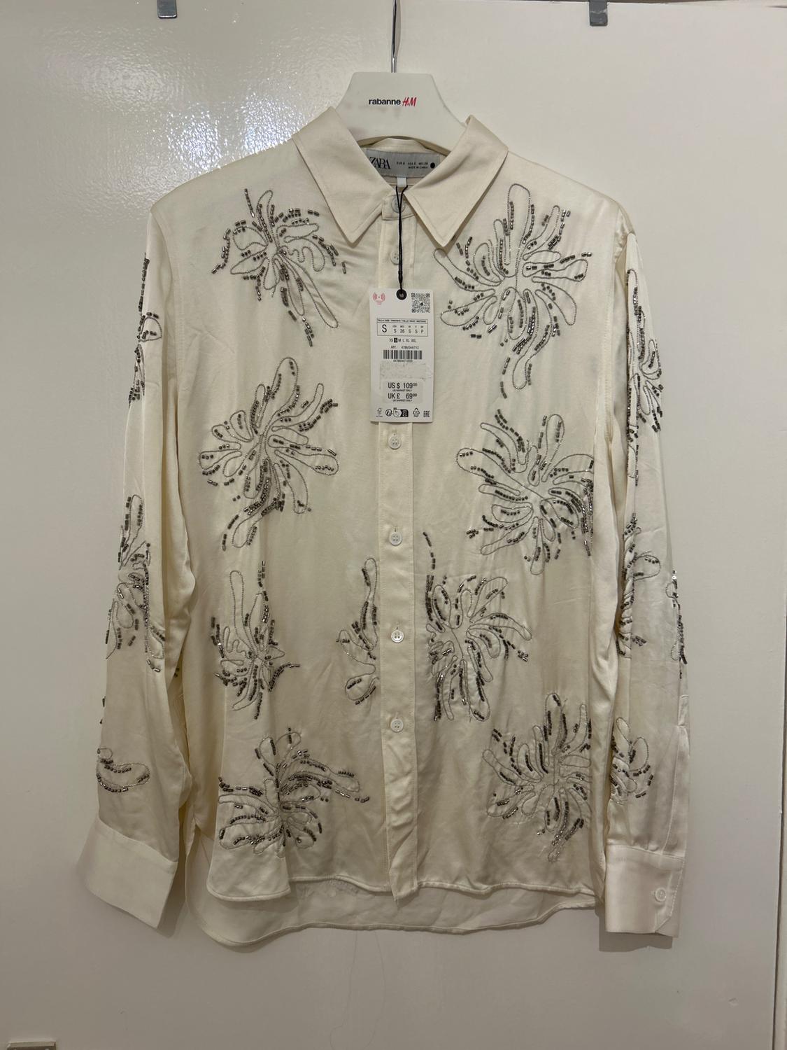 White shirt with black floral embroidery, hanging on a "rabanne H&M" hanger. Visible tag with price and size S.