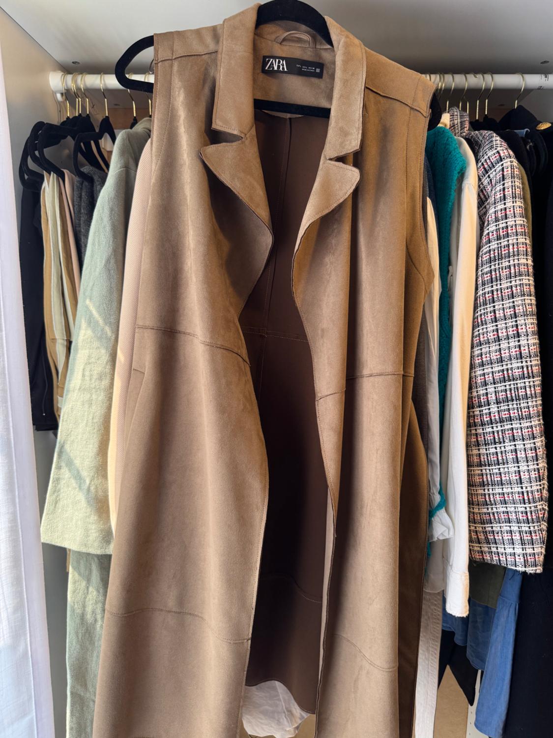 Long brown waistcoat hanging on a hanger inside a wardrobe next to other garments. The label reads ZARA.