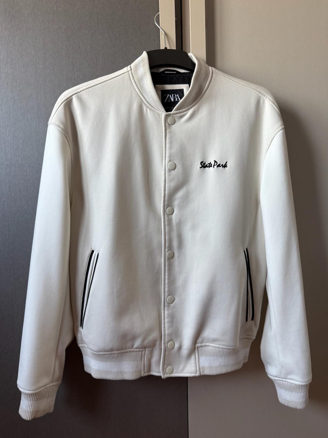 White bomber jacket on a hanger. Press studs, pockets with black trim and "State Park" text on the chest. Visible Zara label.