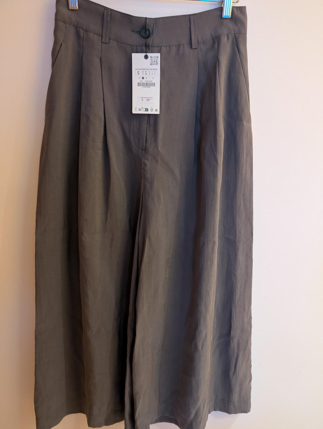 Dark grey wide-leg trousers with pleats and a button, hanging on a hanger against a light background. Label with the text S and € 39.