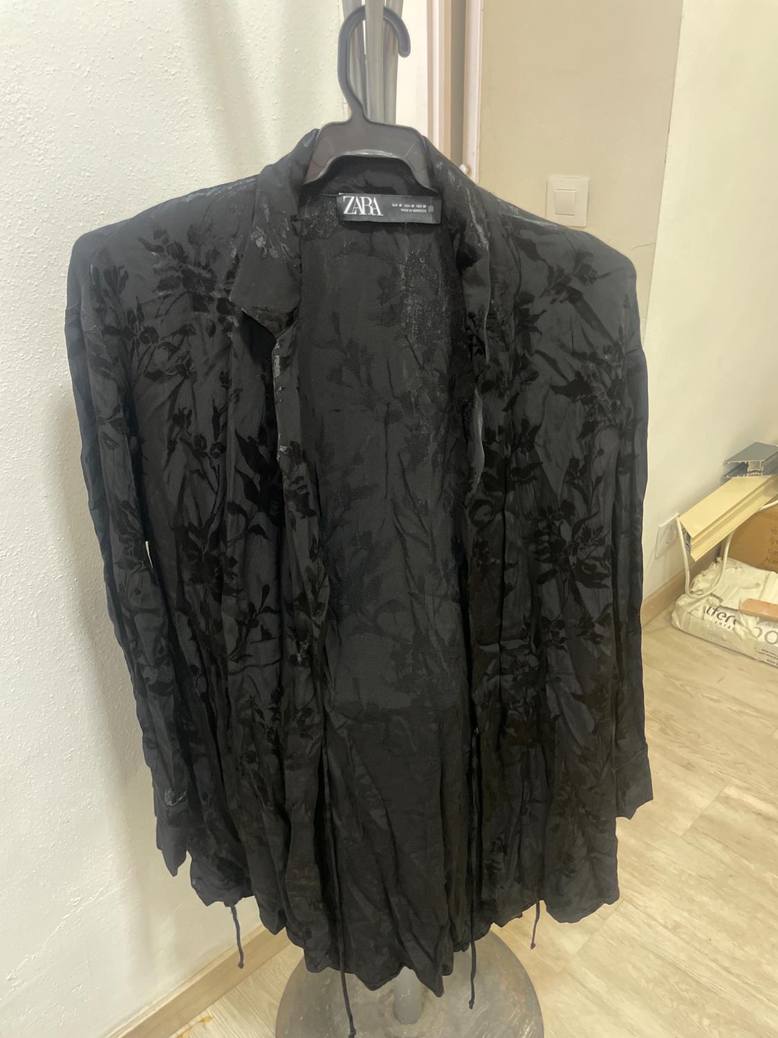 Black jacket with floral print hanging on a clothes hanger. Visible label with the ZARA brand. White wall background and wooden floor.