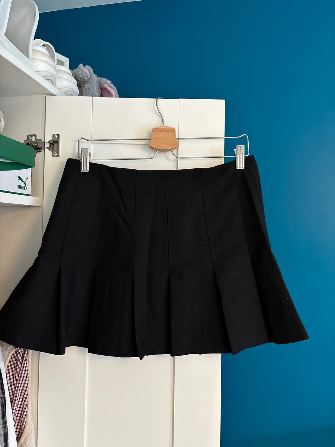 Black pleated skirt hanging on a hanger over a white door. Blue background with shelf displaying trainers and Puma box.