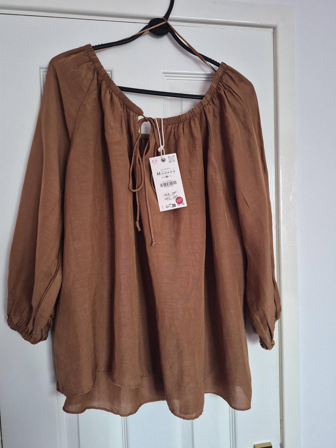 Brown long-sleeved blouse with a gathered neckline and drawstring, hanging on a black hanger against a white door. Label with text “70%”.