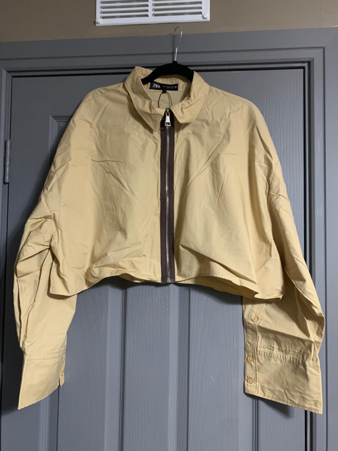 Beige short jacket with front zip and high collar, hanging on a black hanger over a grey door.