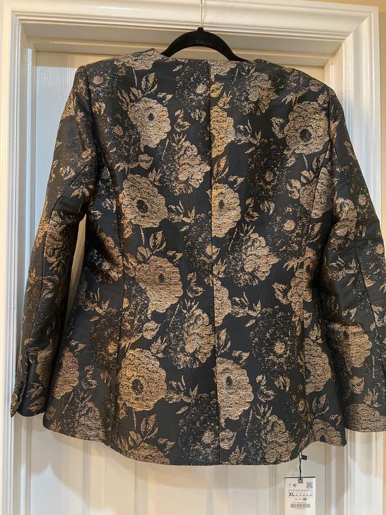 Black jacket with gold floral print, hanging on a hanger. Label visible on the bottom right.