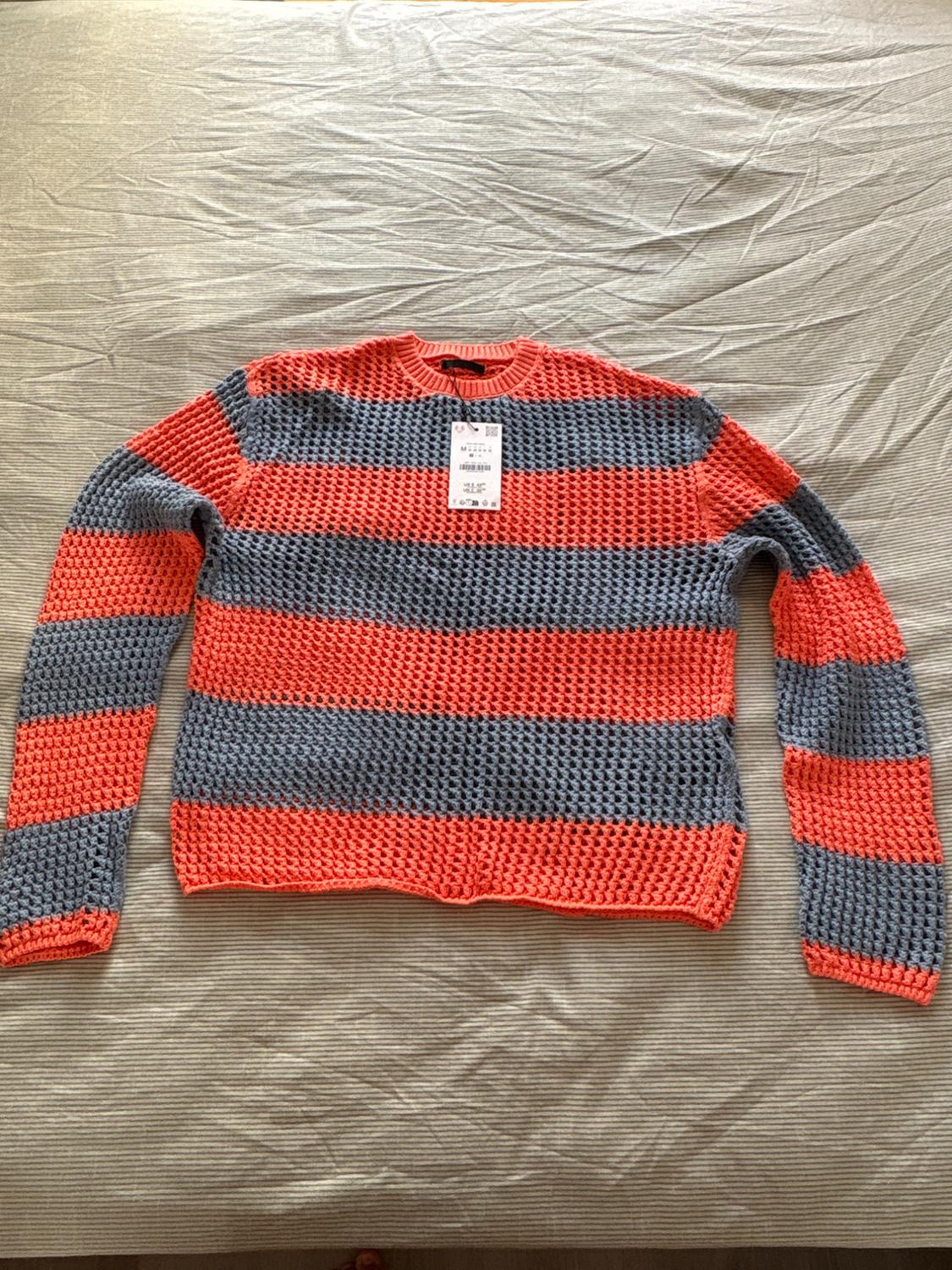 Long-sleeved open-knit jumper with orange and grey stripes. It has a tag attached and is laid out on a bed with striped bed linen.