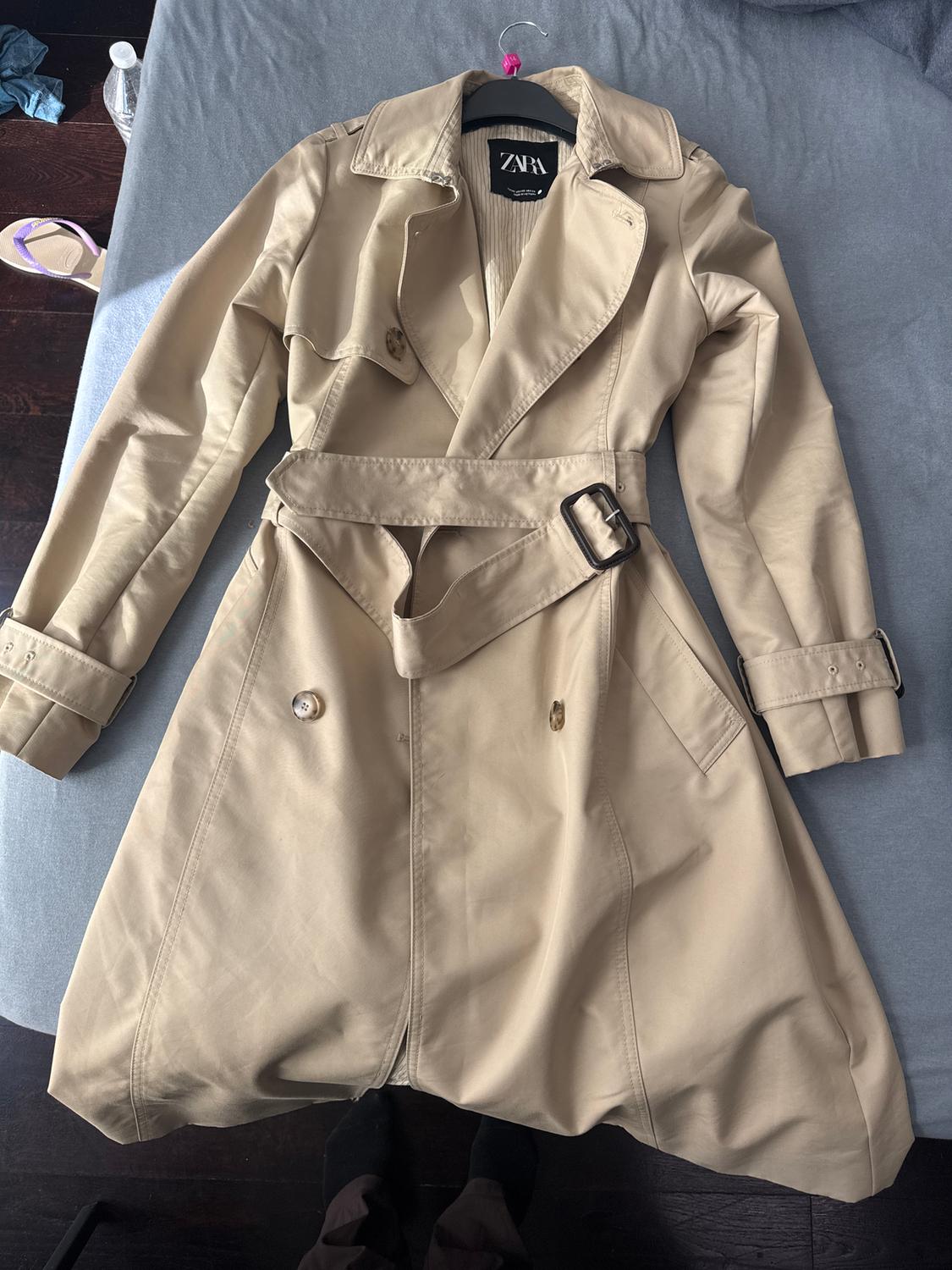 Beige trench coat with a belt and buttons, hanging on a black hanger over a grey bed. Visible label with text "ZARA".