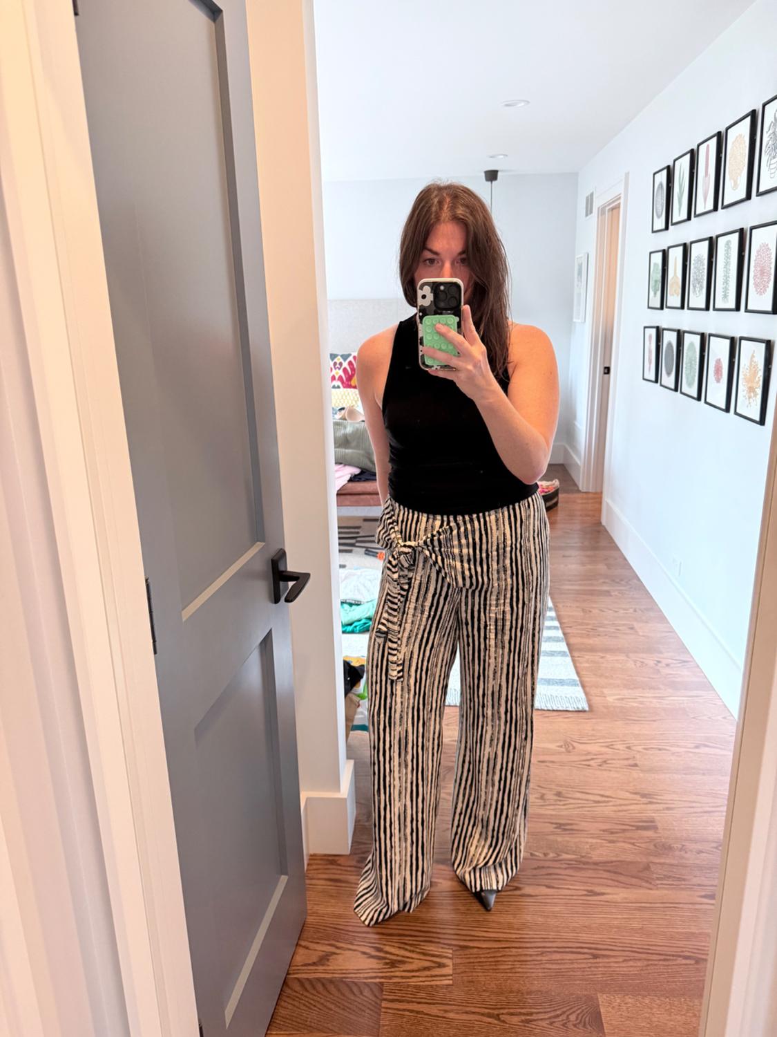 Woman wearing a black top and wide-leg black and white striped trousers, taking a selfie in a mirror in a modern bedroom.