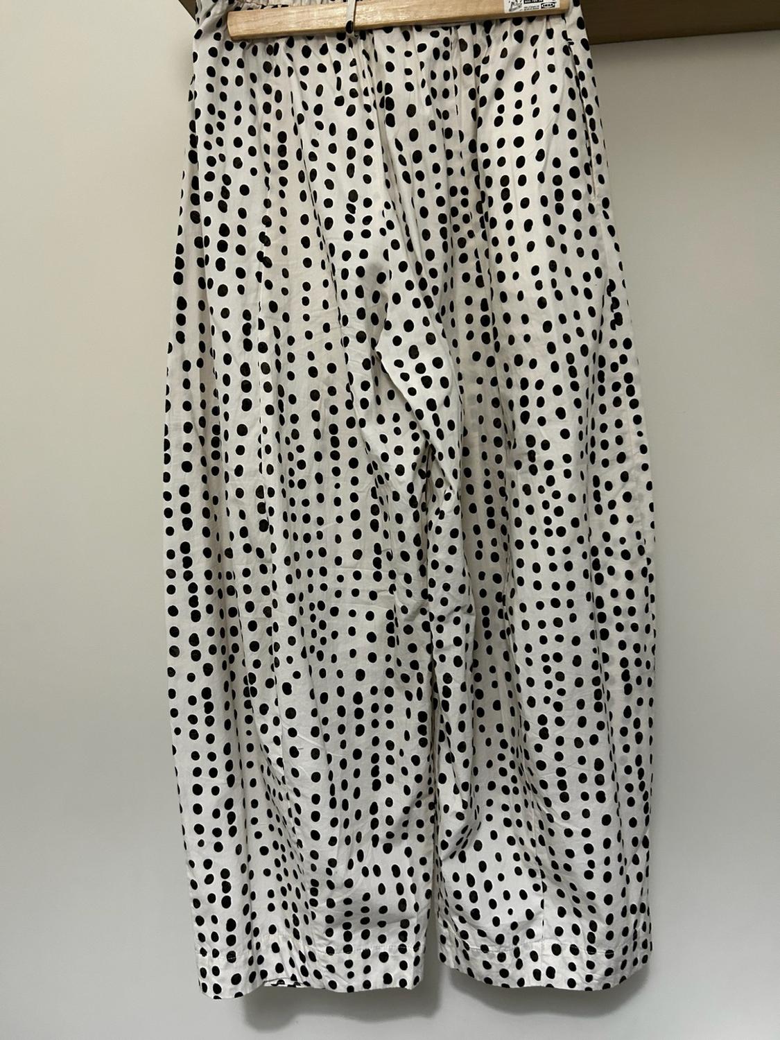 White wide-leg trousers with a black polka-dot pattern hanging on a wooden hanger against a light-coloured wall.