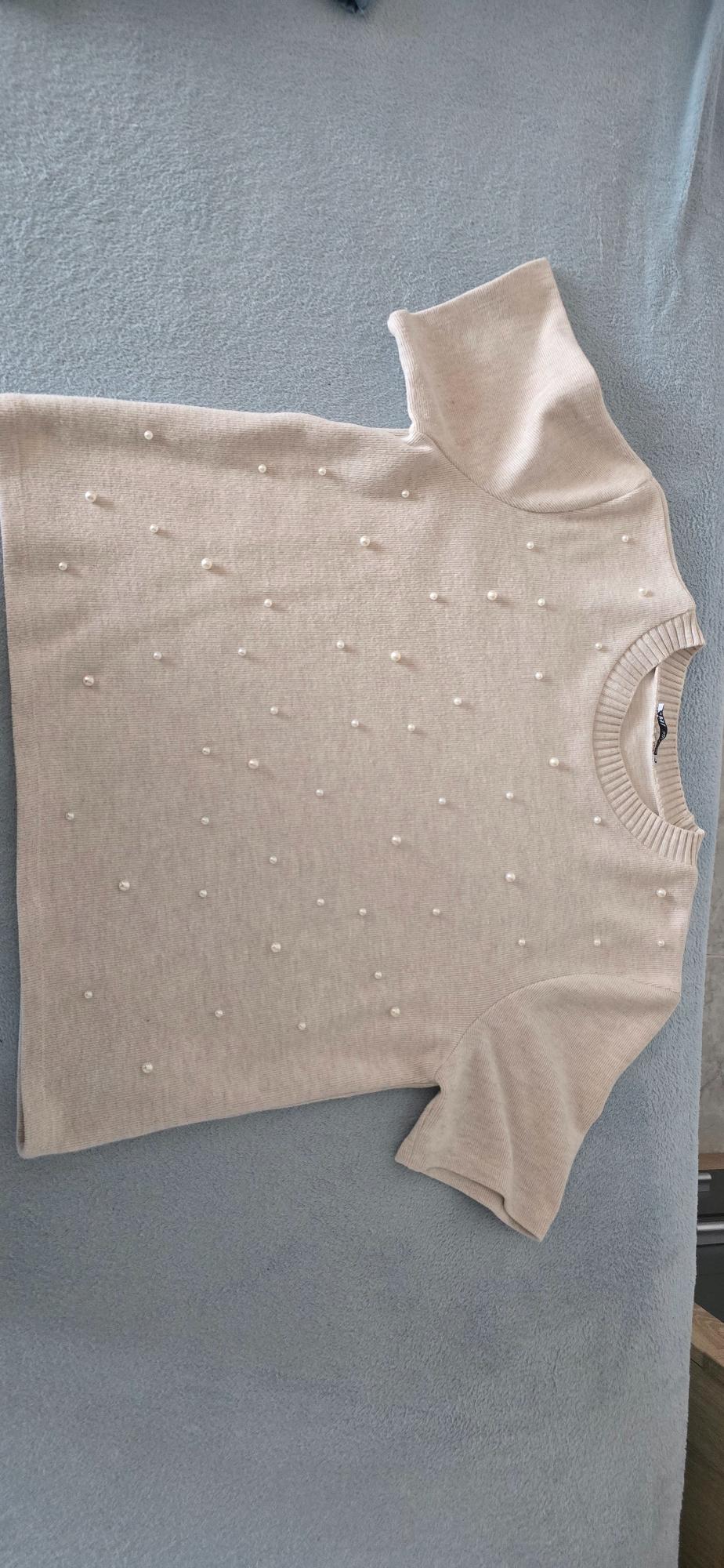 Beige short-sleeved T-shirt with small pearl details, on a grey surface.