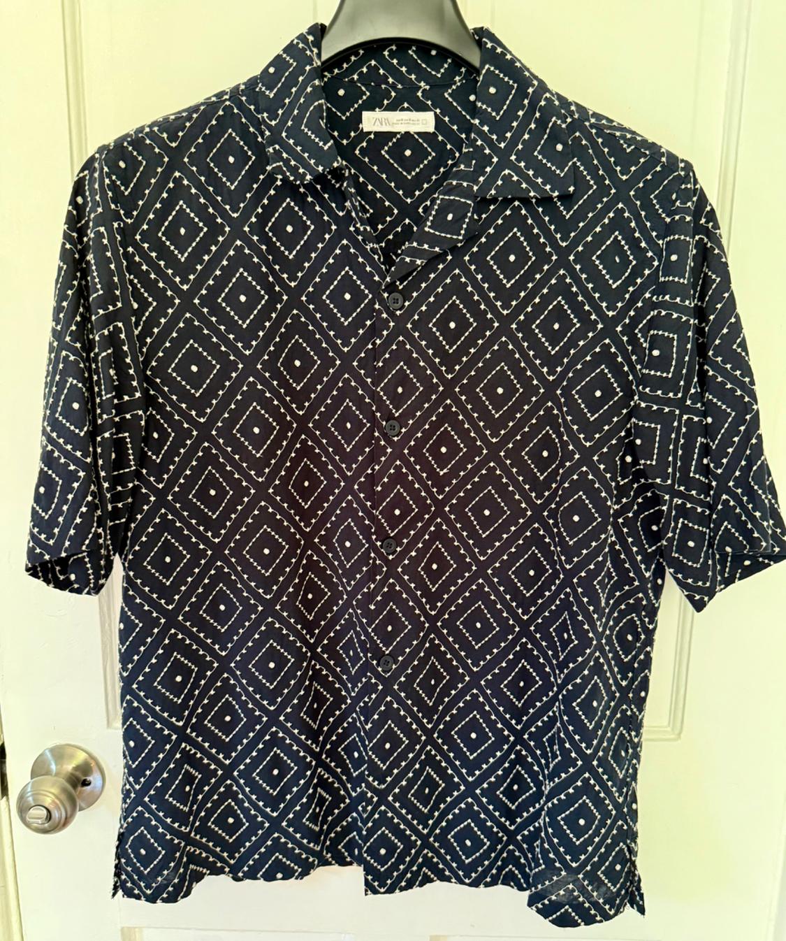 Short-sleeved shirt with a geometric pattern of white diamonds on a black background, hanging on a hanger in front of a white door.
