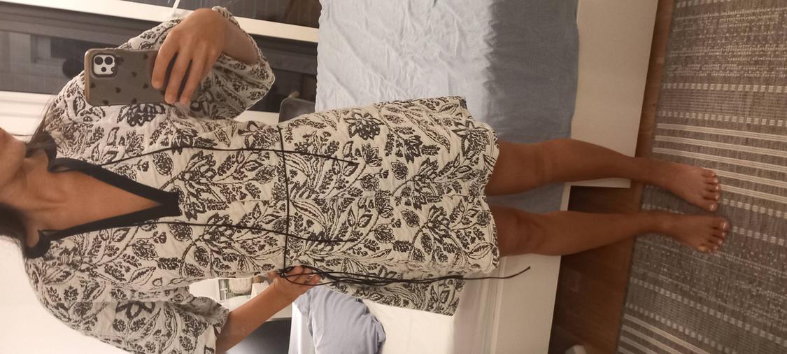 Barefoot woman taking a selfie in her bedroom. She is wearing a short, light-coloured dress with a dark print, a V-neck, and thin ties at the waist.