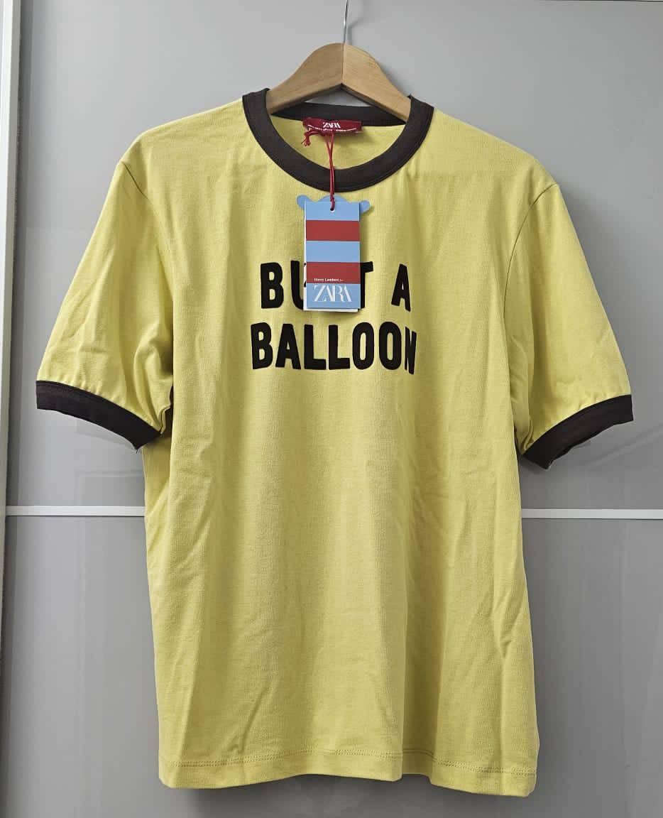 Κίτρινο κοντομάνικο t-shirt με γιακά και σκούρες λεπτομέρειες σε κρεμάστρα. Στο μπροστινό μέρος έχει το κείμενο "BUT A BALLOON".