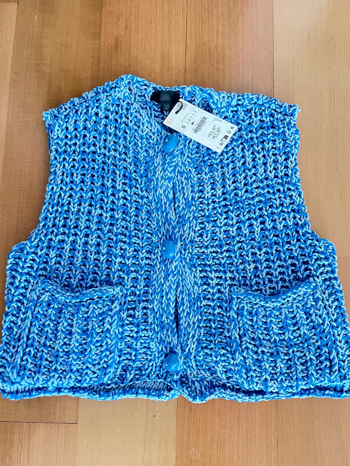 Blue knitted waistcoat with buttons and pockets on a wooden floor. Visible label with size and price details.