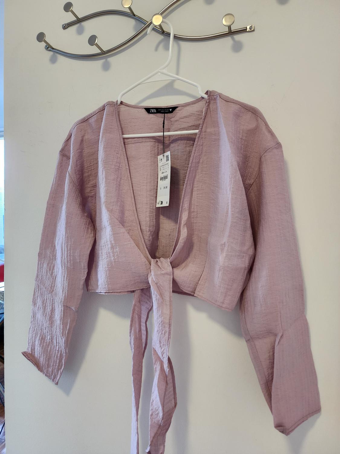 Front view of a pink blouse with a tie at the waist and long sleeves, hanging on a hanger.