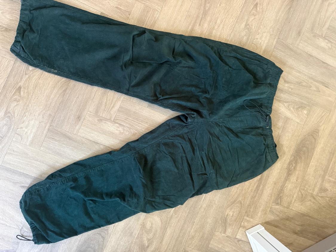 Dark green, loose-fit corduroy trousers with an elasticated waistband and drawstring, laid out on a herringbone wooden floor.