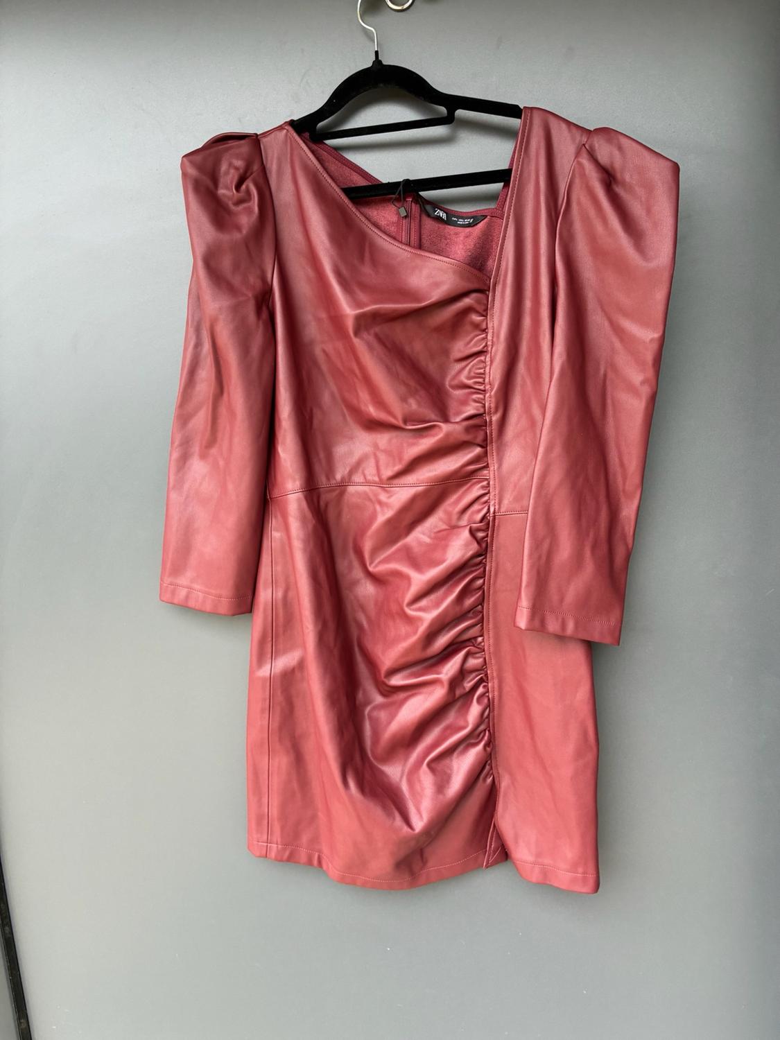 Short burgundy faux-leather dress with puff sleeves and front ruching, hanging on a clothes hanger against a grey background. ZARA label visible.