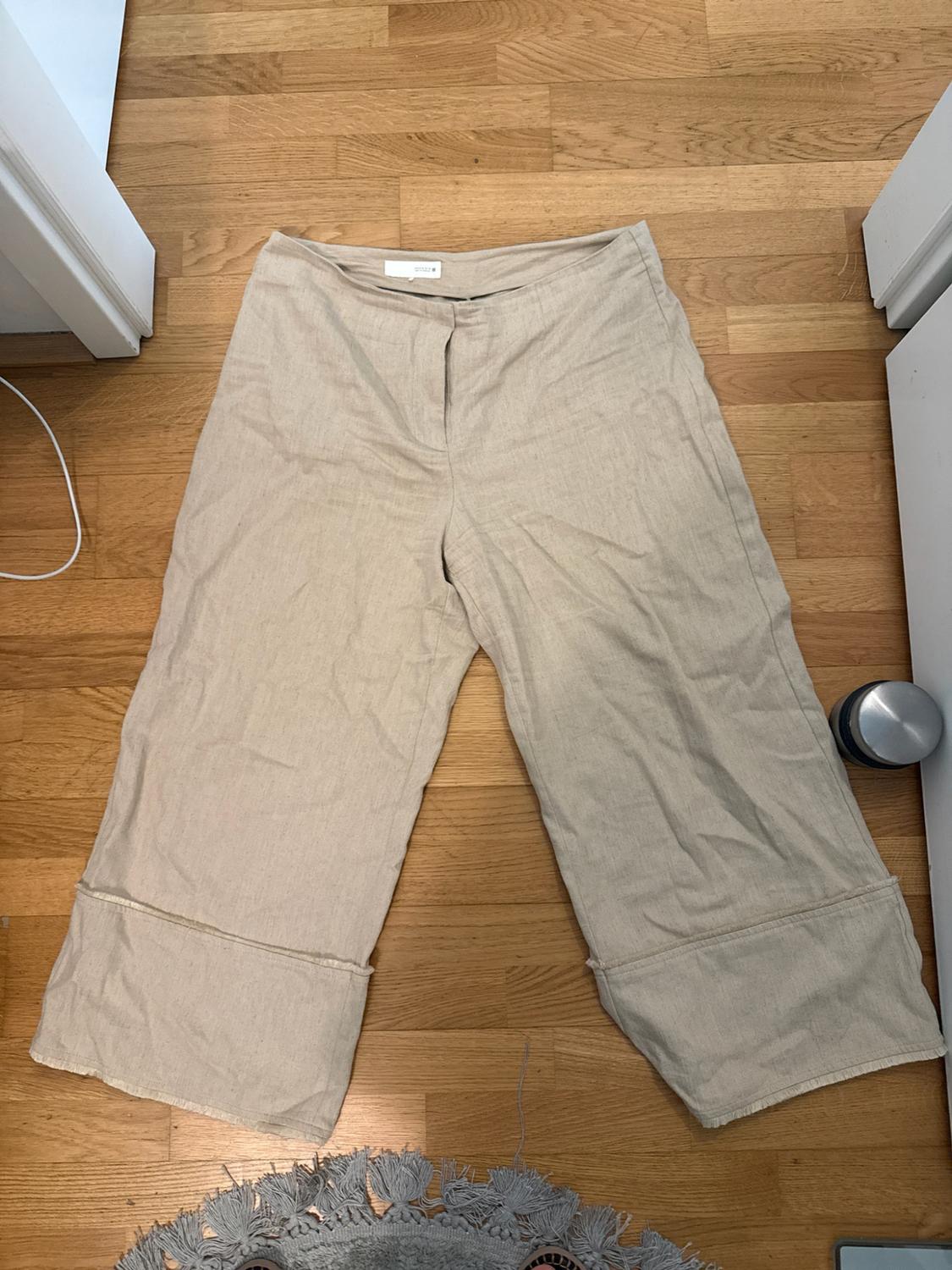 Beige wide-leg trousers with large turn-ups, laid out on a wooden floor next to a grey fringed rug.