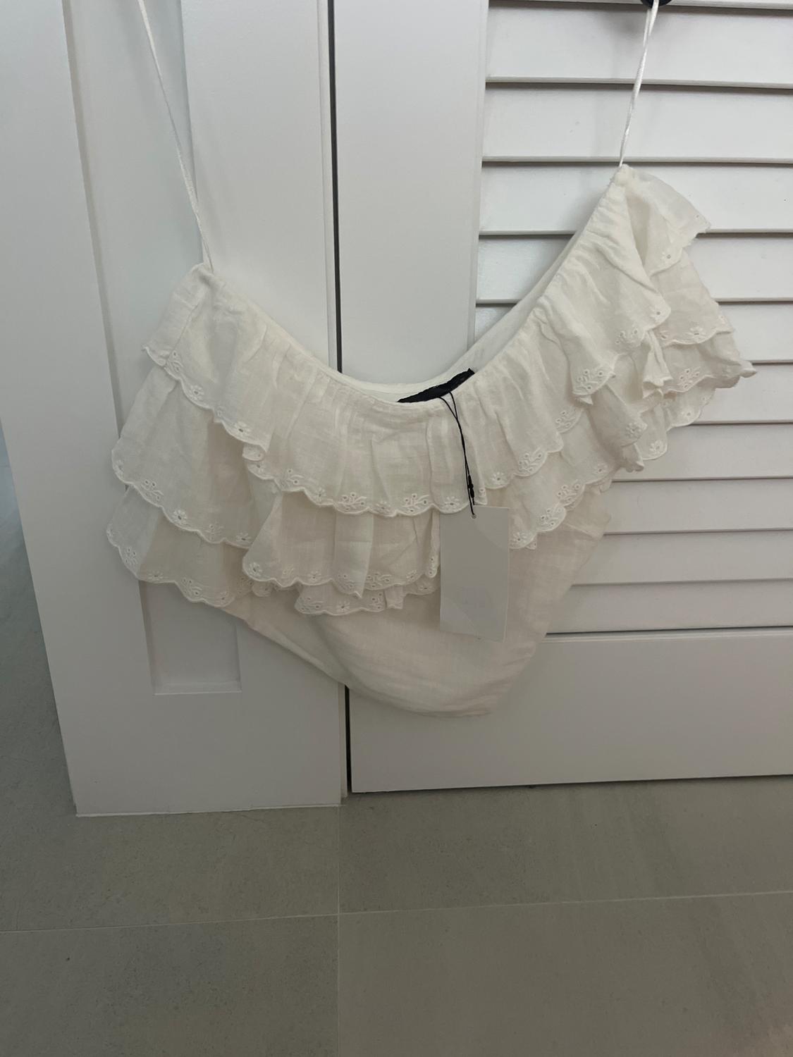 White ruffled lace handbag hanging in front of a white door with blinds. Hanging tag visible.