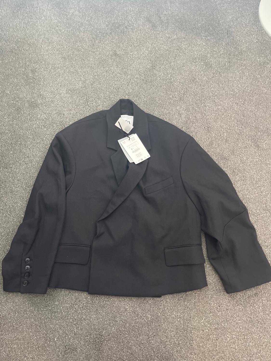 Black jacket with tag on grey carpet. Classic design with lapels and front pockets. Sleeves with buttons.