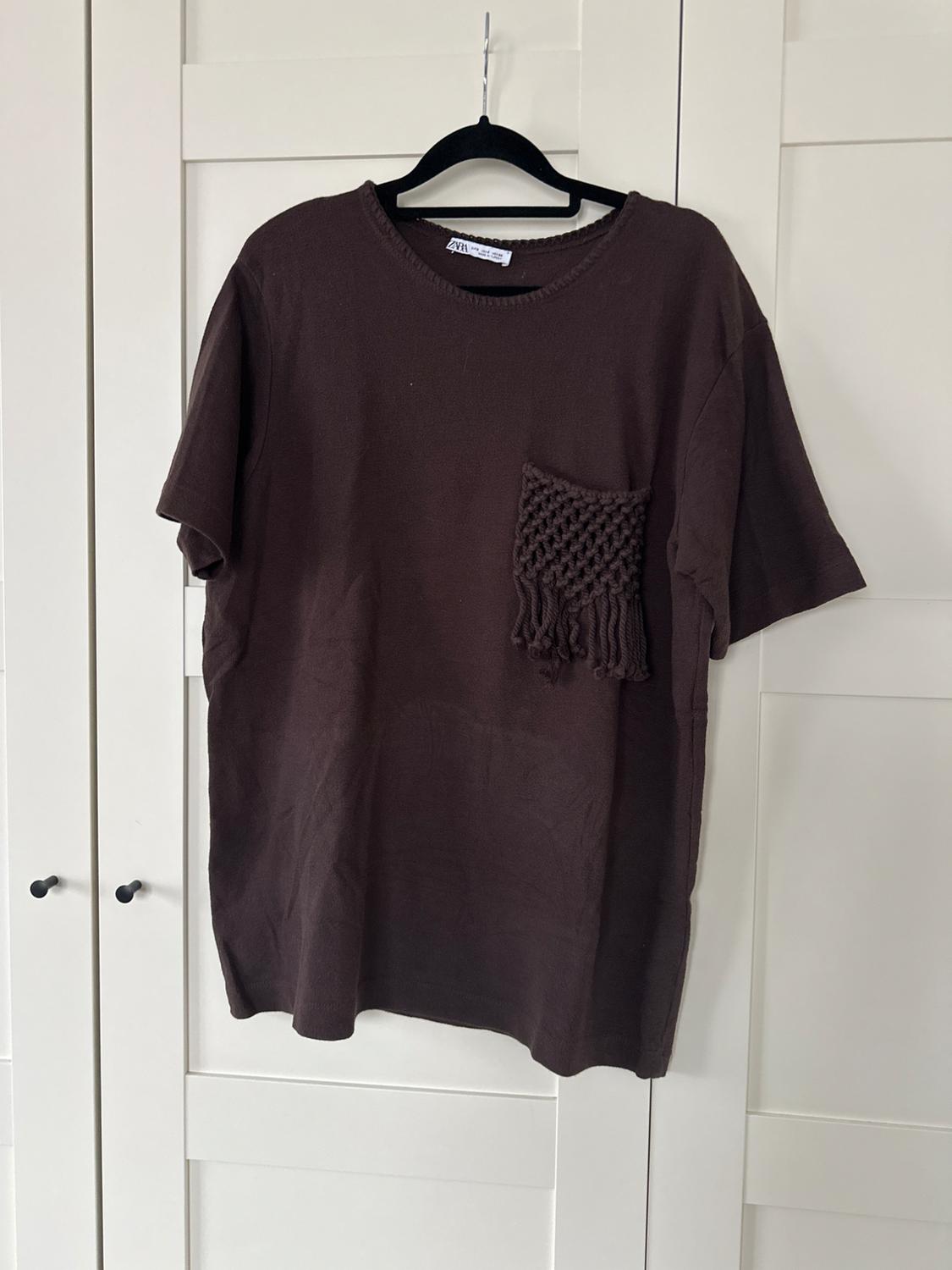 Καφέ σκούρο κοντομάνικο t-shirt zara κρεμασμένο σε μια λευκή ντουλάπα. Ξεχωρίζει μια διχτυωτή τσέπη με κρόσσια στο στήθος.