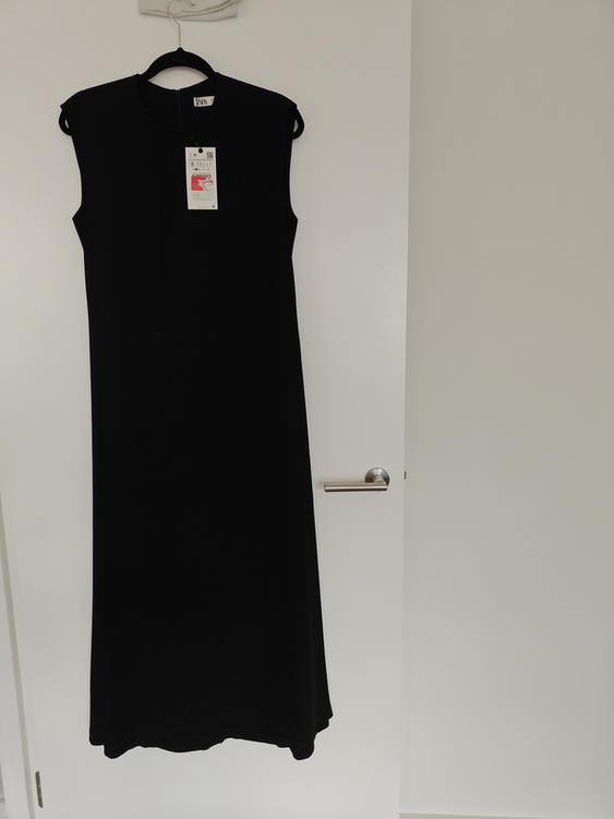 Sleeveless black dress hanging on a white door, with visible tag in the centre of the chest.