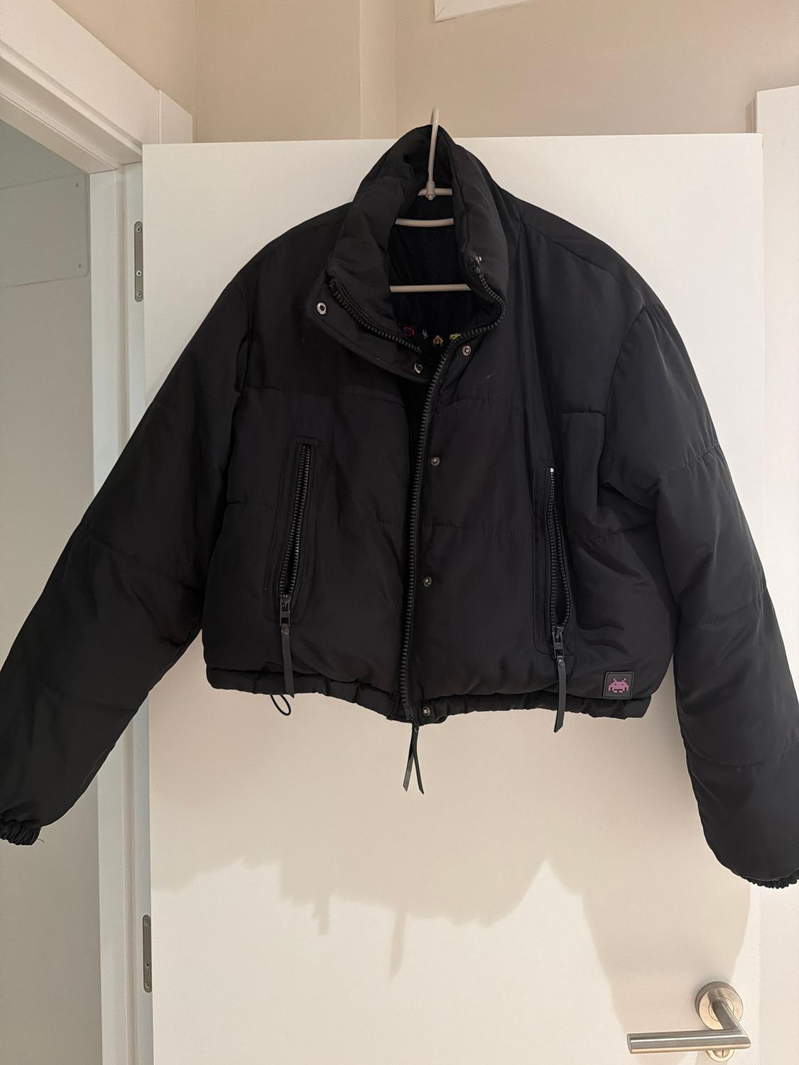 Black padded jacket hanging on a white door. It has a front zip fastening, zip pockets, and a high neck.