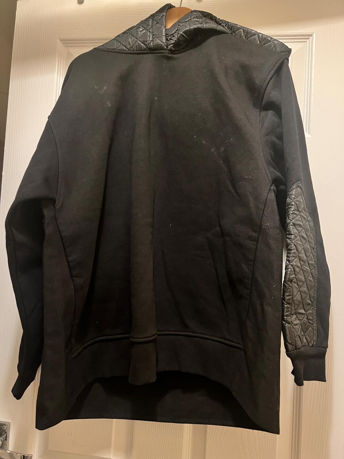 Black hooded sweatshirt with quilted elbow and hood details, hanging on a white door.