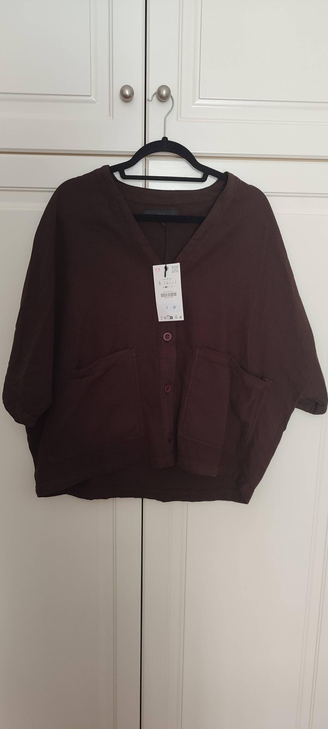 Dark brown jacket with buttons and large pockets, hanging on a black hanger in front of a white wardrobe. Label visible.